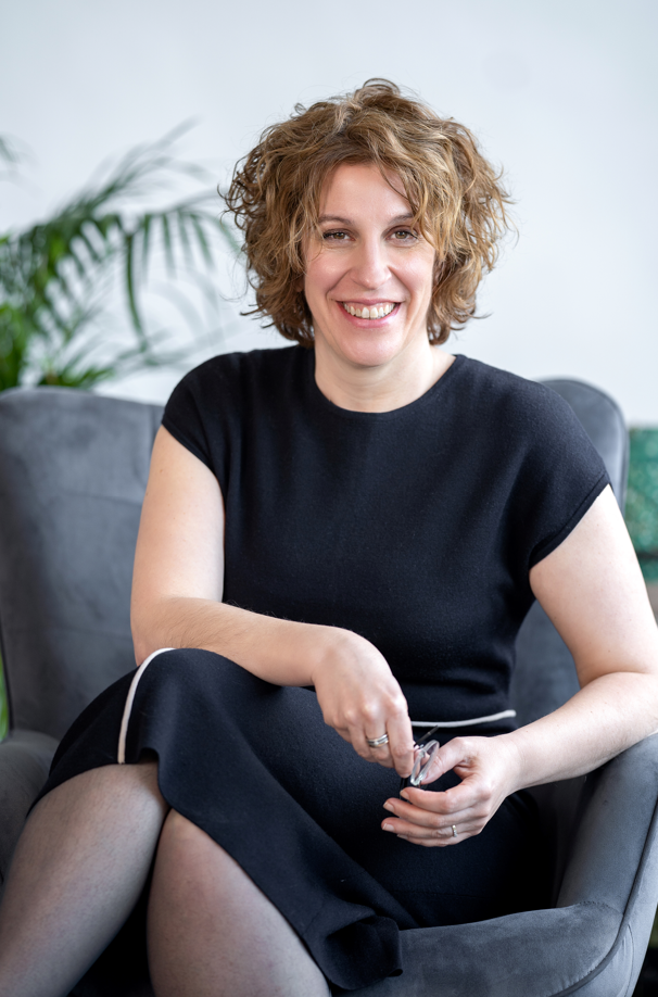 A woman, Gorana Sadnric, formally dressed, sits and smiles to the camera, her hands together.