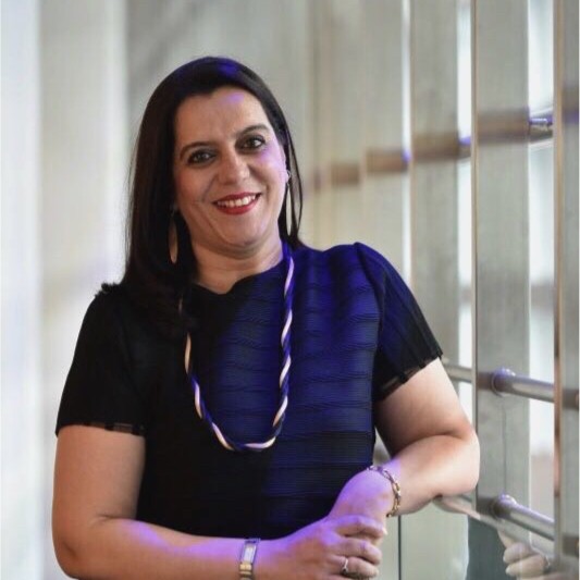 Priya Shahane smiles to the camera, leaning on a glass bannister with her left arm