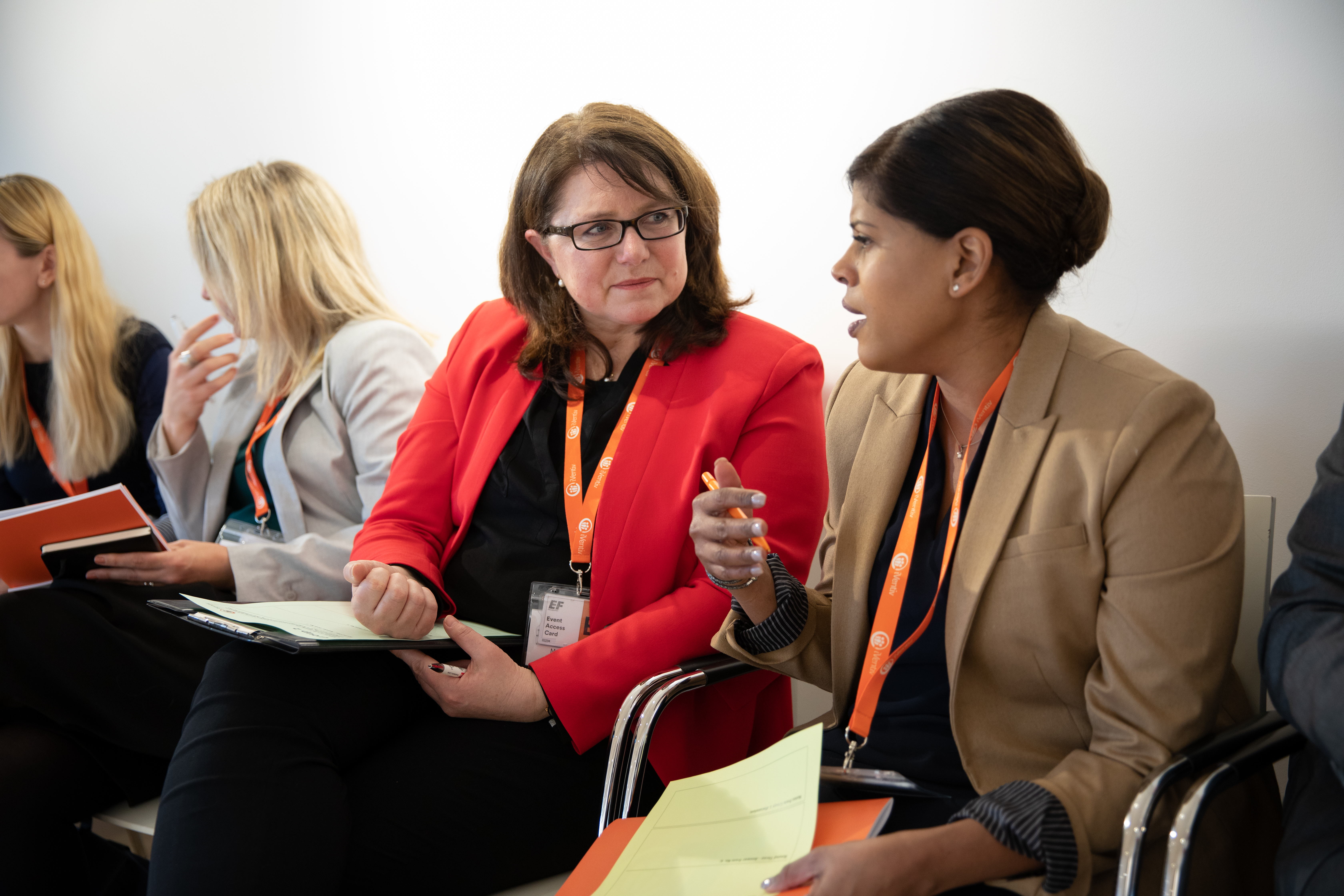 Two women, seated, smile and talk at an executive conference. The woman on the left wears a red jacket and is taking notes.