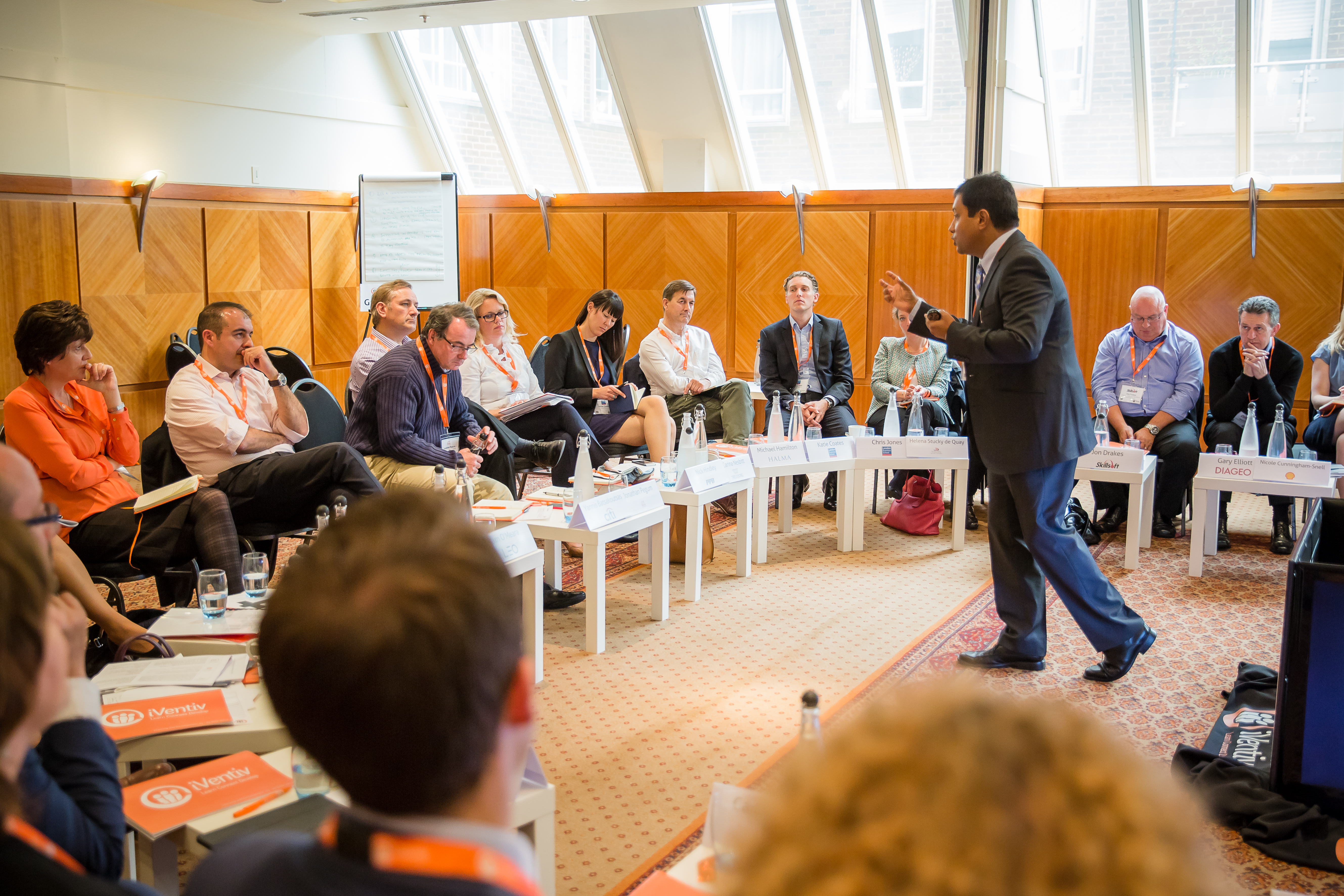 A man stands and strides towards a semi-circle of seated listeners, gesturing forward with his left hand.